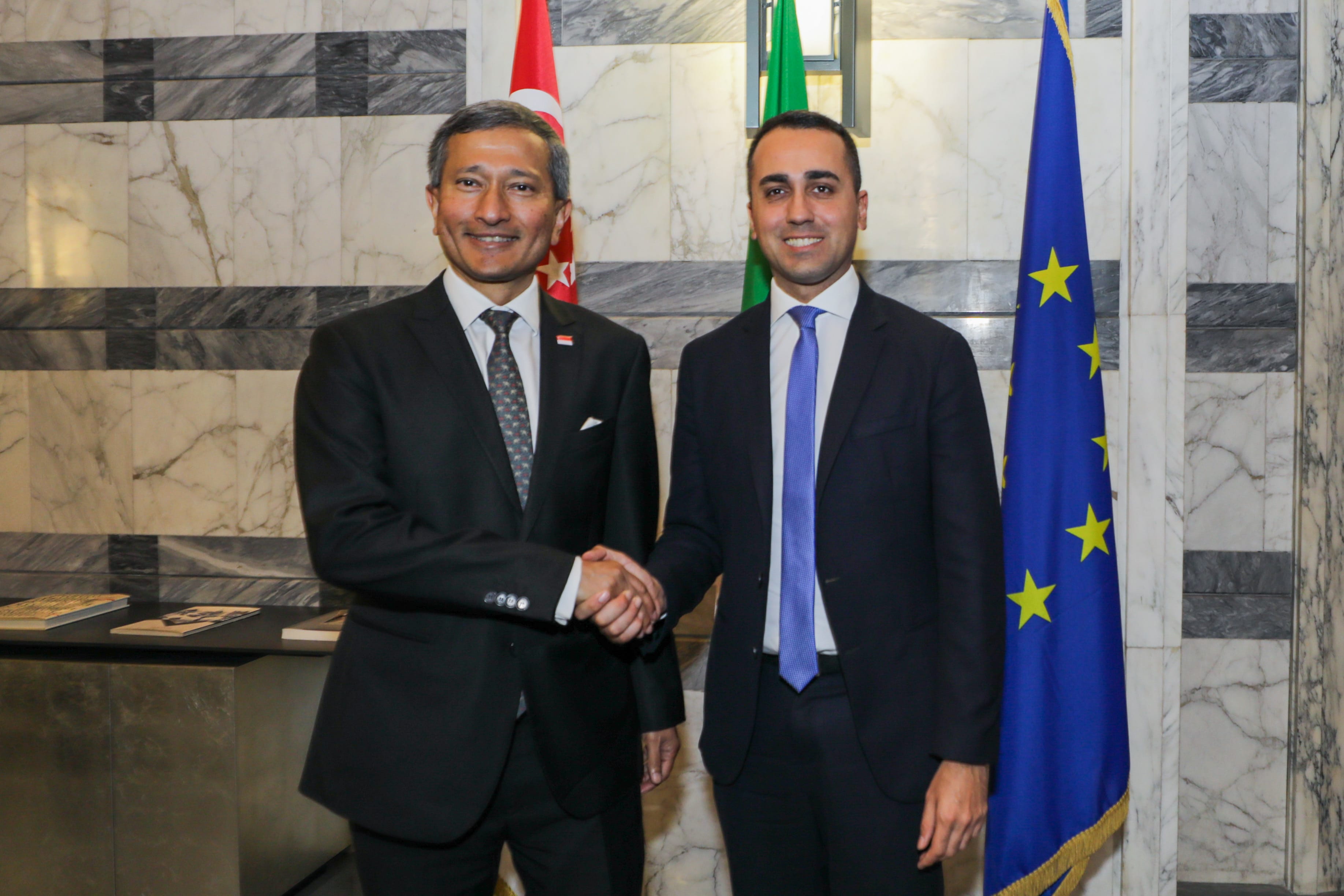 Two men in suits shake hands near flags of Singapore, Italy, and the EU.